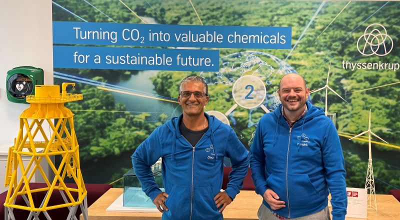 Zwei Carbon2Chem®-Mitarbeiter am Messestand mit der Wand „Turning CO₂ into valuable chemicals“